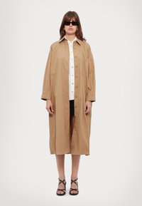 Giacca camicia oversize marrone con colletto, caratterizzata da chiusura con bottoni, una tasca, indossata sopra una camicia bianca e pantaloni corti neri. Sandali neri con cinturini.