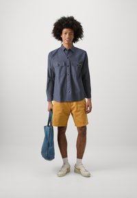 Button-up shirt in navy with two chest pockets; paired with mustard shorts. Carrying a blue tote bag, wearing white sneakers and socks.