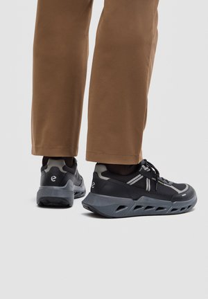Persona che indossa pantaloni marroni e scarpe sportive nere e grigie con suola spessa, vista da dietro su uno sfondo chiaro e uniforme.