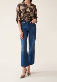 Chemise à motifs floraux marron avec des manches retroussées, associée à des jeans évasés bleus et des chaussures à talons pointus beiges. Une simple ceinture noire souligne la taille.