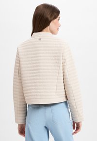 Beige gewatteerde jas met een cropped ontwerp, hoge kraag en subtiele logo-details op de achterkant, gecombineerd met lichtblauwe denim jeans.