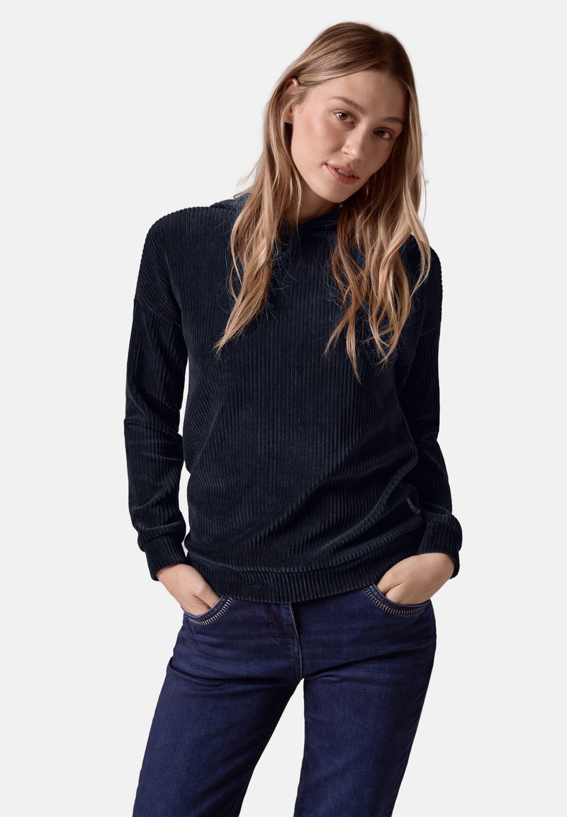 Maglietta a maniche lunghe a coste di colore blu scuro, con una vestibilità rilassata e scollatura rotonda. Abbinata a jeans scuri con dettagli di cucitura discreti.