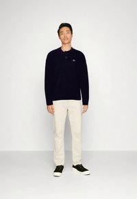 Marineblå polotrøye med langermet og krage, med en liten logo. Beige bukser og svarte joggesko kompletterer antrekket. Ensfarget bakgrunn.