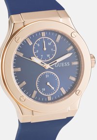 Reloj Round Guess con un reloj azul y marcadores de hora en tono dorado, doble subdial y correa de silicona azul. Caja de acero inoxidable dorado.