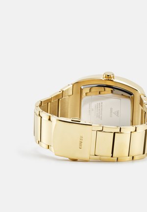Montre en métal doré avec un boîtier rectangulaire, un bracelet en acier inoxydable et une finition polie ; présente un logo de marque visible sur le fermoir.