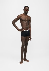 Moški model stoji boso, oblečen v črne Moschino boksarice na preprostem belem ozadju, ena roka na boku.