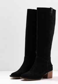 Black suede knee-high boots with a rounded toe, a stacked wooden heel, and a side zipper. Features simple stitching and a logo accent.