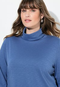 Modrý rolák z měkké, strukturované látky. Má přeložený límec a volný střih, doplněný stříbrnými kroužkovými náušnicemi.