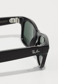 Occhiali da sole neri in plastica con lenti verdi, caratterizzati da una forma rettangolare piatta e una cerniera in argento. Logo Ray-Ban visibile sul lato.