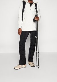 Giacca impermeabile bianca, pantaloni neri e scarpe da trekking beige. Porta bastoni da trekking. Zaino con spallacci visibili.