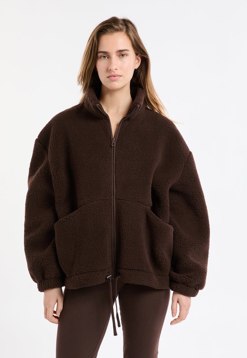 Brązowa kurtka polarowa z wysokim kołnierzem, zamkiem błyskawicznym, dużymi przednimi kieszeniami i regulowanym dolnym trokiem. Miękka tekstura i luźny krój.