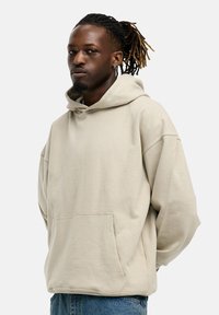 Beige Kapuzenpullover mit Kängurutasche, oversized Schnitt, strukturiertem Stoff, großer Kapuze und gesenkten Schultern. Darunter werden Jeans aus Denim getragen.