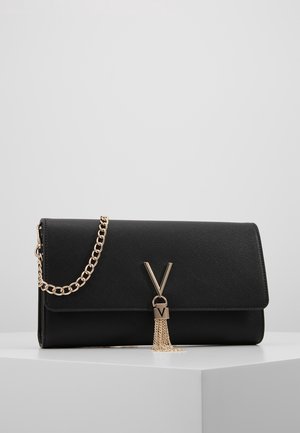 Zwarte gestructureerde clutch met gouden kettingriem en decoratieve gouden "V" met kwastje op het voorflapje, geplaatst op een witte ondergrond.