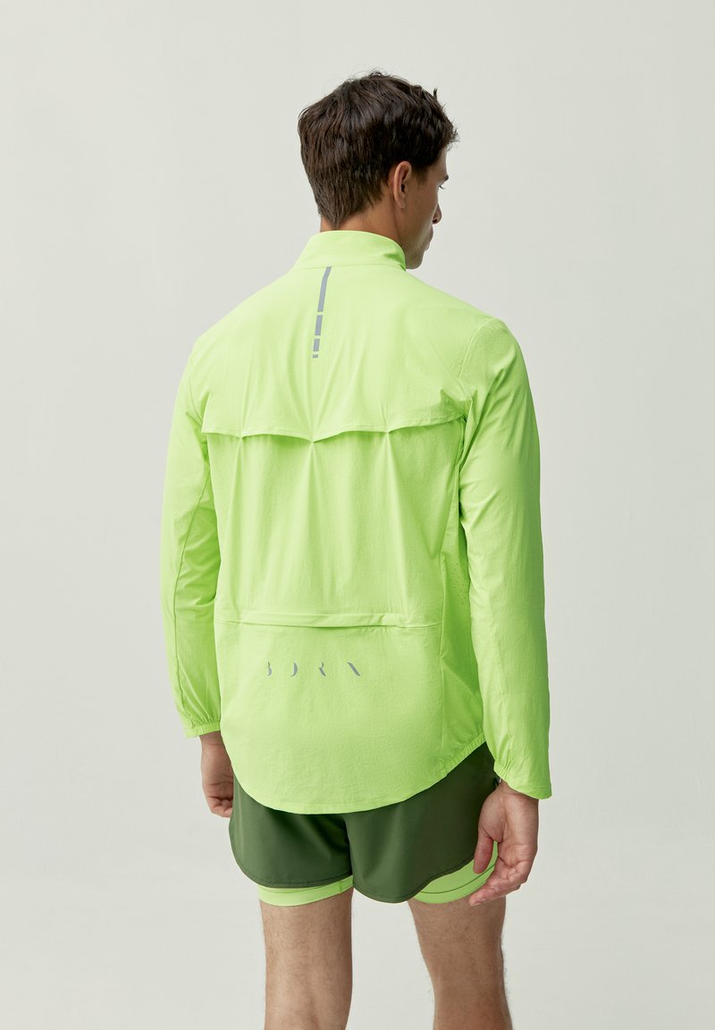 Giacca leggera di un vivace verde lime con maniche lunghe, aperture posteriori e dettagli riflettenti. Abbinata a pantaloni corti verde oliva.