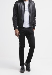 Chaqueta bomber de cuero negra con puños acanalados, llevada sobre una camiseta blanca. Combinada con jeans negros ajustados y zapatos negros brillantes con cordones.