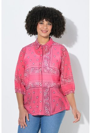 Uśmiechnięta kobieta o kręconych włosach, ubrana w różową koszulę na guziki z wzorem i rękawami do łokcia oraz niebieskie dżinsy, na tle białego tła.