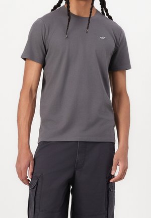 Mand iført en ensfarvet grå t-shirt med korte ærmer og sorte cargoshorts, stående mod en hvid baggrund.