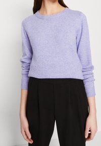 Mujer vestida con un suéter de cuello redondo lavanda con puños acanalados y pantalones de cintura alta plisados negros, de pie contra un fondo liso.