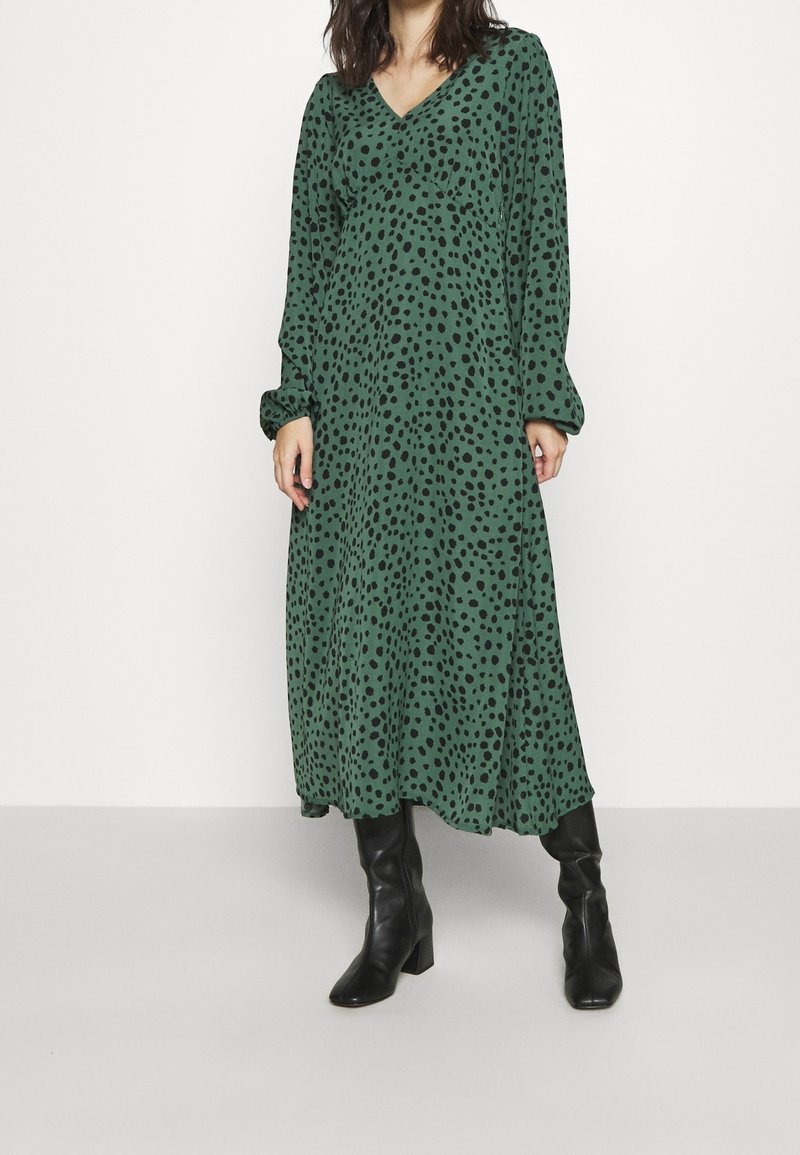 Grünes Midi-Kleid mit schwarzen Punkten, V-Ausschnitt, langen Ärmeln und lockerer Passform. Getragen mit kniehohen schwarzen Stiefeln. Glatte Textur.