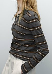 Polo gris à manches longues avec des rayures horizontales marron et blanches. Il présente un col et des poignets côtelés, associé à un pantalon blanc.