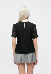 Μαύρο τοπ από δαντέλα με κοντά μανίκια, με οδοντωτές άκρες και λεπτομέρεια κλειδώματος στην πλάτη. Φοριέται με μια ανοιχτό γκρι πλισέ φούστα.