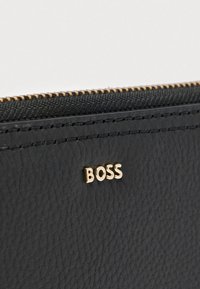 Suprafață texturată din piele neagră cu fermoar auriu și litere mici aurii care formează cuvântul „BOSS” aproape de marginea superioară.