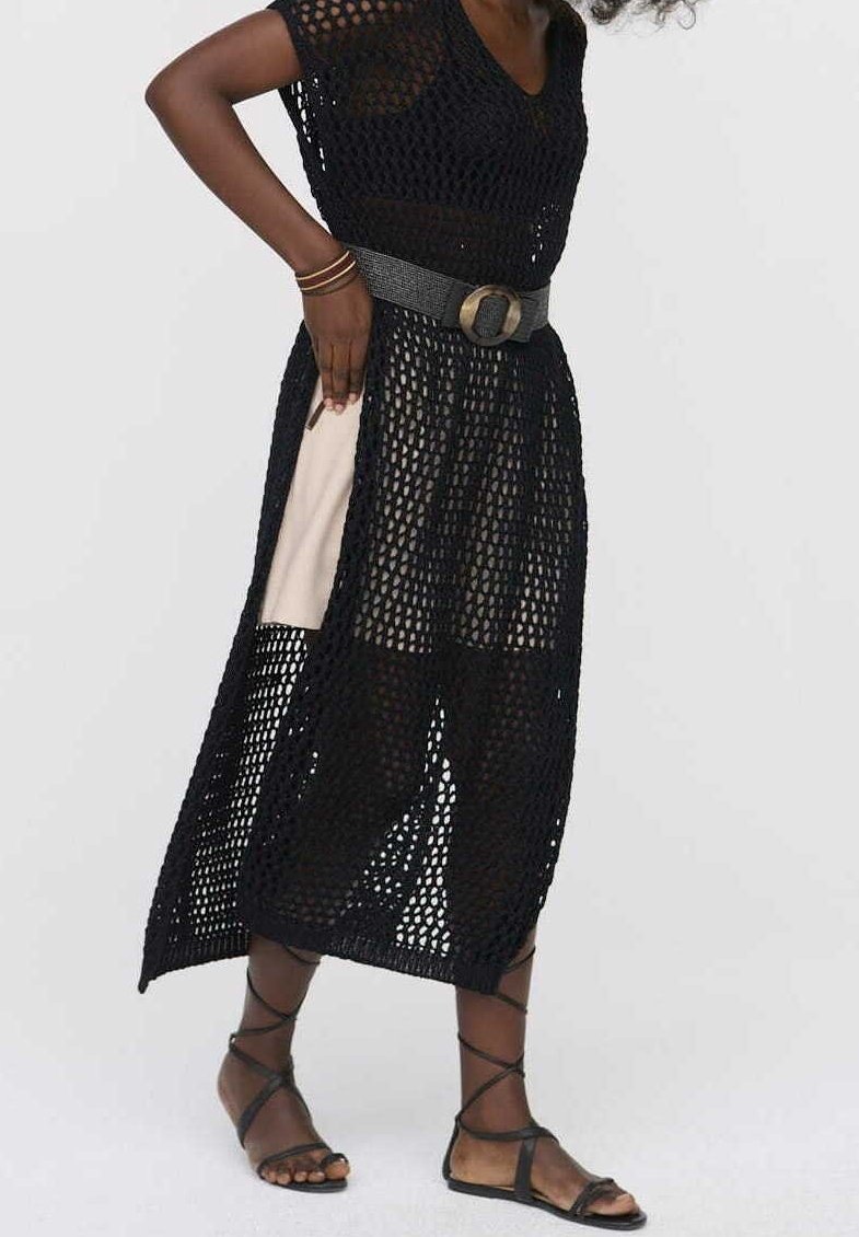 Femme portant une robe noire en crochet par-dessus un short beige, accessoirisée avec une ceinture large, des bracelets et des sandales noires à brides.