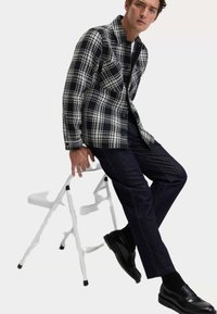 Czarna i biała koszula w kratę z guzikami, noszona na białej koszulce, w zestawieniu z ciemnoniebieskimi dżinsami i czarnymi loafersami, siedząca na stołku.