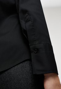 Camicia da dress nera con colletto, caratterizzata da una texture liscia, con polsino a bottoni e vestibilità sagomata. Dettaglio della manica in primo piano su uno sfondo grigio.