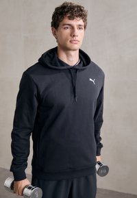Ung mand i sort Puma hoodie, der holder 6 kg krom vægte, stående foran en ensfarvet lysvæg.