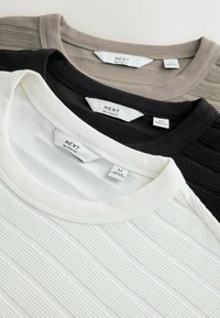 Trois chemises à col rond côtelées en beige, noir et blanc, chacune étiquetée "NEXT Relaxed Fit" avec les étiquettes de taille visibles.