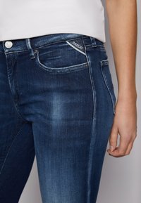 Jeans de mezclilla azul oscuro con efecto de desgaste, cinco bolsillos y herrajes en plata. Presenta una etiqueta de logo blanca cerca de la cinturilla.