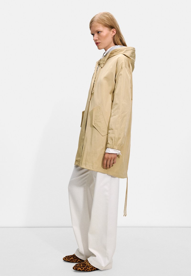 Veste légère beige avec capuche, deux poches avant, ourlet à cordon de serrage, portée avec un pantalon large blanc et des chaussons imprimés léopard.