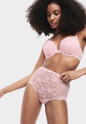 Donna che aggiusta la lingerie di pizzo rosa a vita alta, indossando un reggiseno abbinato, con capelli ricci e voluminosi, su uno sfondo chiaro e semplice.