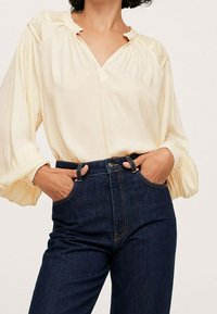 Blusa leggera color crema con scollo a V e colletto arricciato, abbinata a jeans in denim blu scuro a vita alta.