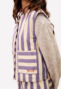 Gestreifter Weste in lila und cremigem Baumwollstoff, mit einer Seitentasche und besticktem "Buchfink". Getragen über einem strukturierten beigen Pullover.