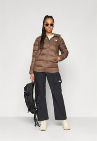 Ruda pūkinė striukė su užtrauktuku priekyje, juodos darbo kelnės, balta viršutinė dalis ir smėlio spalvos batai; modelis laiko juodą kuprinę, dėvi baltus akinius nuo saulės.