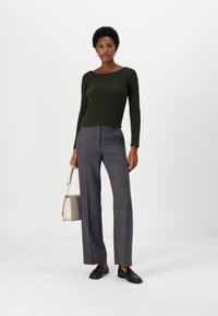 Pull en maille côtelée vert foncé, pantalon gris à carreaux bien ajusté, mocassins noirs et un petit sac à main beige. Le modèle se tient contre un fond blanc.