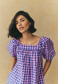 Lila- und rosa Karodress mit Puffärmeln, quadratischem Ausschnitt und gerafften Details. Weicher, strukturierter Stoff mit seitlichen Taschen.