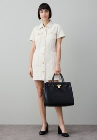 Een crèmekleurige blousejurk met korte mouwen; gecombineerd met een zwarte leren handtas met gouden accenten en een gestructureerde vorm.
