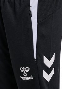 Pantalon de sport noir avec des panneaux latéraux blancs, une poche zippée, deux chevrons blancs et un logo blanc hummel près de la poche.