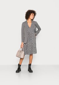 Blazer gris sur une robe noire à motifs avec une taille cintrée. Des bottines noires et un sac à main beige clair complètent la tenue.
