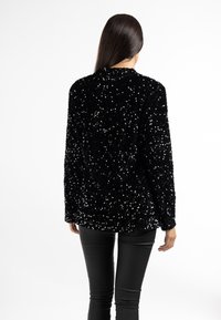 Femme aux longs cheveux noirs portant une veste noire à paillettes et un pantalon noir, tournée de dos sur un fond blanc.