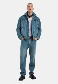 Giacca di jeans blu e jeans, entrambi con una texture consumata. Maglietta bianca sotto, scarpe nere e una sciarpa nera attorno al collo.