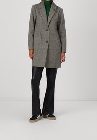 Abrigo gris a cuadros con cuello de muesca, cierre de dos botones, grandes bolsillos, llevado sobre un suéter de cuello alto verde, combinado con pantalones de cuero sintético negros y zapatillas.