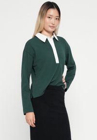 Polo vert à manches longues avec un col blanc et une patte de boutonnage. Le tissu semble lisse avec une coupe décontractée, associé à une jupe noire.