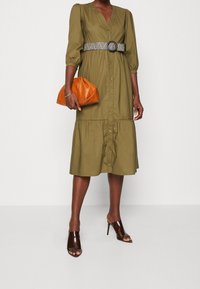 Zielona oliwkowa sukienka z guzikami i warstwowym dołem, z czarno-białym plecionym paskiem, zestawiona z pomarańczową kopertówką i brązowymi sandałami na obcasie.