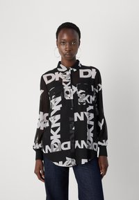 Svart gjennomsiktig skjorte med store hvite DKNY-logoer, knappelukking foran, to brystlommer og elastiske mansjetter, kombinert med mørke jeans.