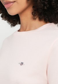 Camiseta de algodón rosa claro con cuello redondo y mangas cortas. Presenta un pequeño logo bordado en la zona del pecho. Textura suave.