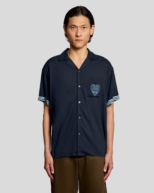 Mand iført en marineblå kortærmet skjorte med knapper og "Clyde's Laundry" hjerte-logo på brystlommen samt brune bukser.